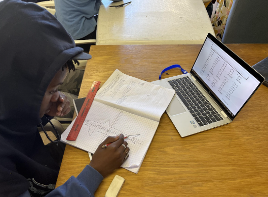A boy in a hoodie sitting at a table, engaged in educational activities on his laptop. Zoe Education Trust
