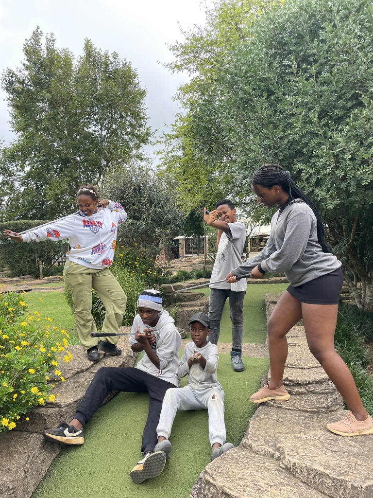 Four people of varying ages enjoying a playful moment together at an outdoor setting with greenery in the background. Zoe Education Trust