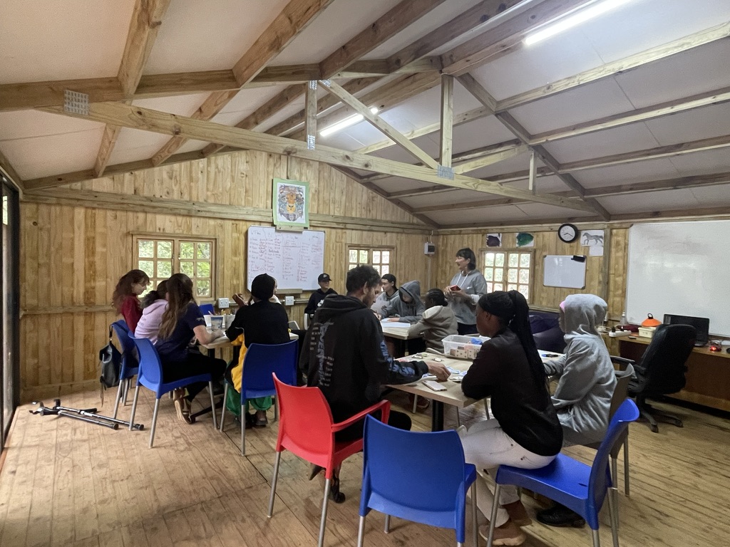 A group of individuals engaged in a meeting or class inside a wooden cabin with colorful chairs. Zoe Education Trust