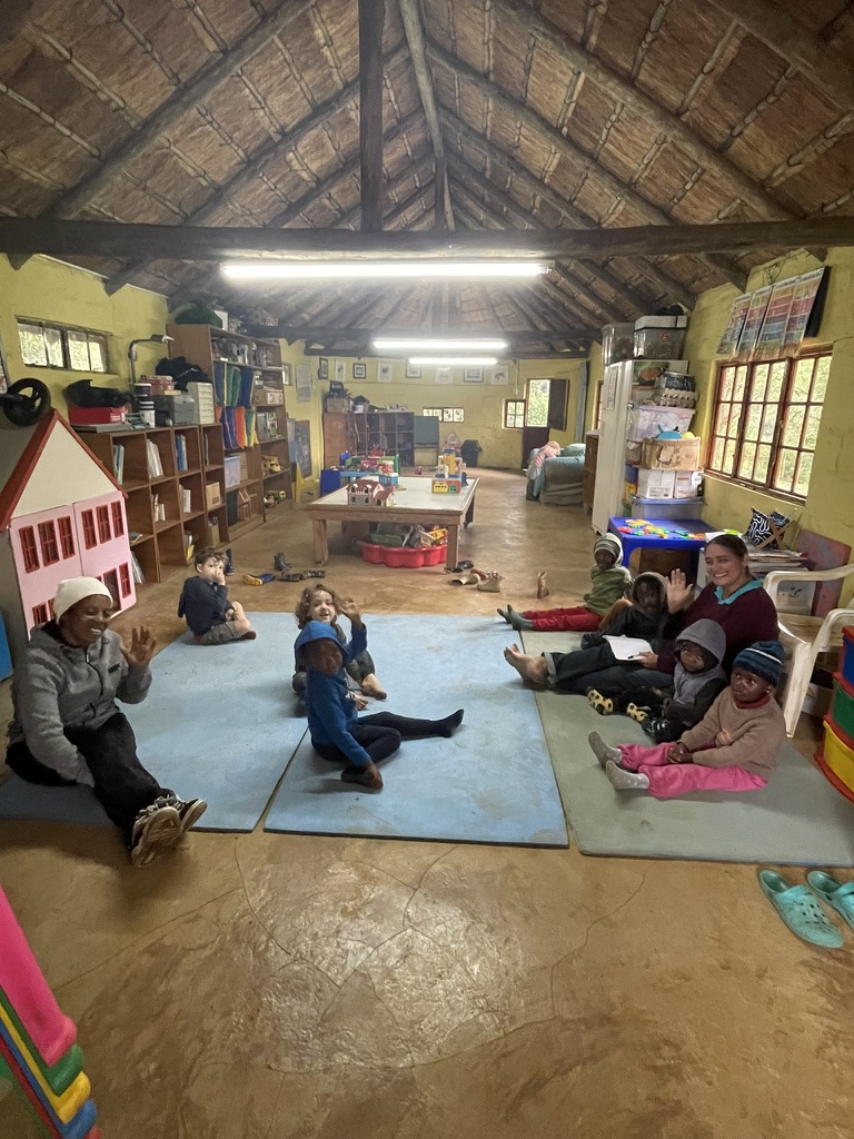 A group of children and adults sitting on the floor inside a cozy, well-stocked preschool classroom. Zoe Education Trust