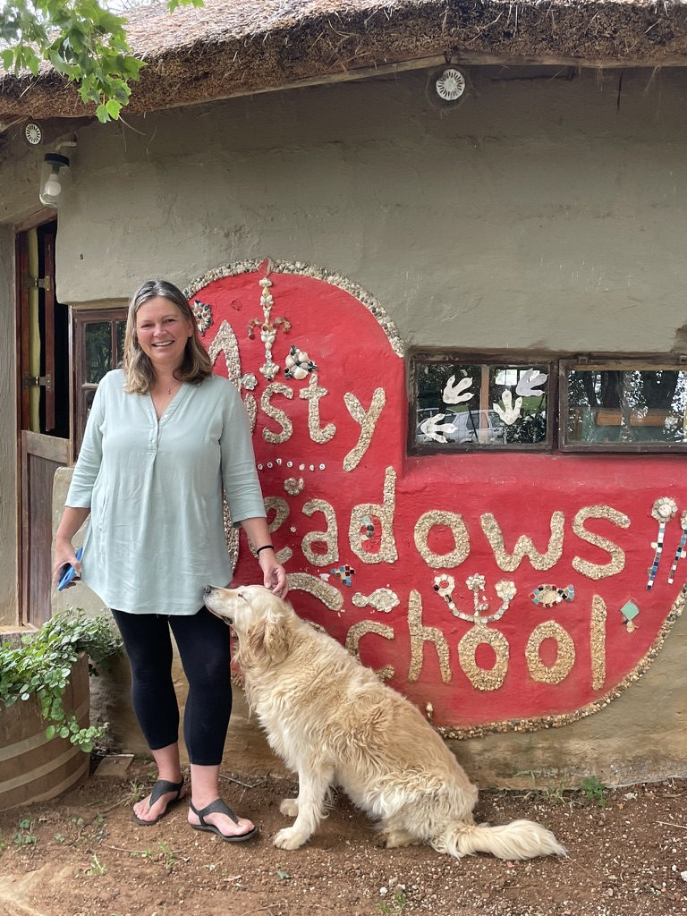 A smiling woman standing next to a golden retriever in front of a building sign that reads "misty meadows school. Zoe Education Trust