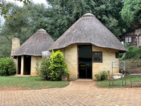 Traditional thatched-roof huts with a paved walkway in a lush green setting. Zoe Education Trust